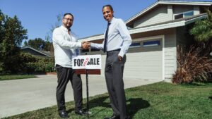 Two men shaking hands in front of a large home with a “for sale” sign and a “sold” sticker on top of it.
